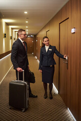 Business traveler with luggage being assisted by hotel staff in hallway. Hotel employee opening...