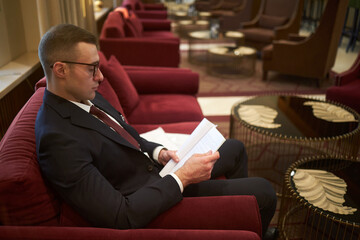 Business professional examining papers while sitting on red couch in modern lounge with...
