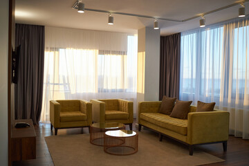 Modern living room with yellow furniture, sheer curtains, and a warm ambiance created by ample natural light flowing through large windows, highlighting elegant decor