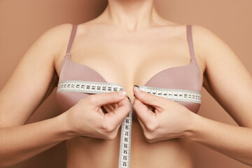 Woman in beautiful bra measuring breast circumference on beige background, closeup