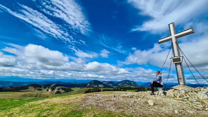 Hiker pauses near summit cross on Plankogel, Styria, Austria. Panoramic view of rolling green hills...