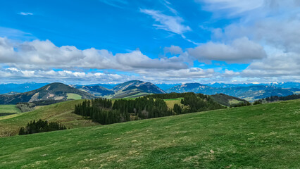 Fototapeta premium Verdant rolling hills across Plankogel, Styria, Austria, under partly cloudy sky. Patches of coniferous trees dot the landscape, creating a textured, scenic view of the Austrian countryside. Tranquil