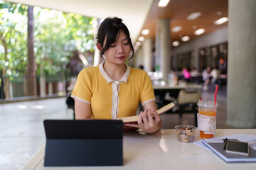Close-up of a woman's hands writing and reading a book on a study desk with a laptop and drink