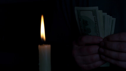 A person holds a few banknotes while standing next to a flickering candle. The soft glow of the candle contrasts with the shadows in the room, creating a mysterious atmosphere.