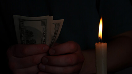 Two hands grip a few hundred dollar bills, illuminated by the warm glow of a candle, casting shadows in a dimly lit environment, creating a cozy yet mysterious atmosphere.