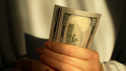 A person clasps a hundred dollar bill with both hands, showcasing its details. The soft light creates a warm atmosphere as the bill is held against a blurred background.