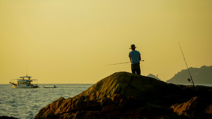A fisherman stands on a large rock, casting his fishing line into the calm sea during sunset. Nearby, boats float peacefully, creating a serene atmosphere by the water.