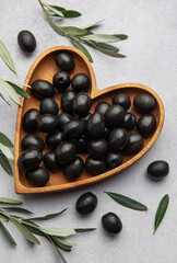 Black olives arranged in heart shaped wooden bowl on gray background with olive branches