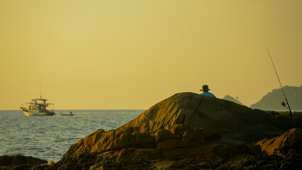 A person sits on a large rock by the water, fishing peacefully as a boat glides across the calm sea during a beautiful sunset in a serene coastal location.