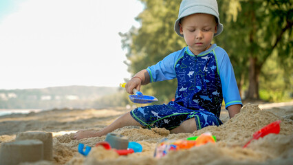 A young child sits on the sand at a beach, focused on playing with colorful toys. The scene is bright and sunny, surrounded by lush trees and calm water.