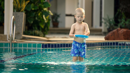 A young child stands in shallow water, holding a blue frisbee while wearing patterned swim shorts. Bright sunlight illuminates the cheerful atmosphere around the pool.