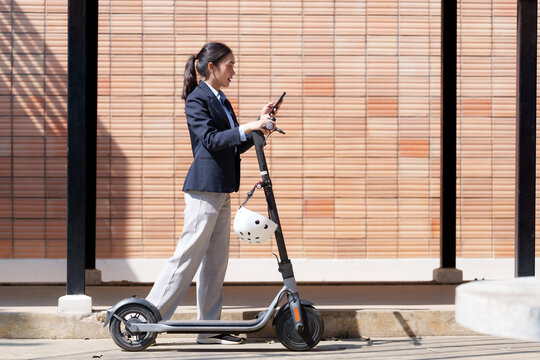 Businesswoman in a suit is using her smartphone while riding an electric scooter, showcasing modern commuting and technology integration in an urban environment