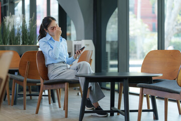 Stressed businesswoman rubbing her tired eyes while using smartphone in office lobby, suffering...