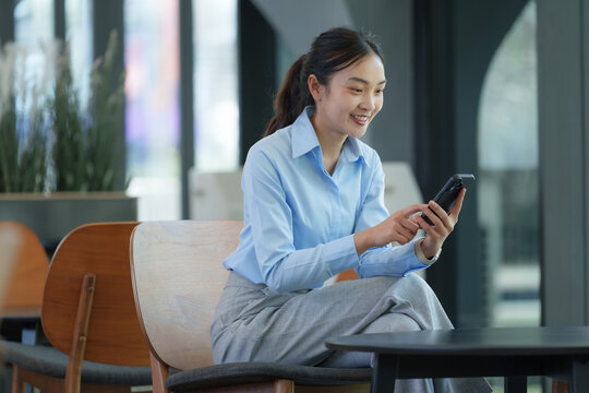 Young asian businesswoman sitting on a chair in a modern office, using her smartphone with a smile, showcasing the concept of mobile technology in the workplace