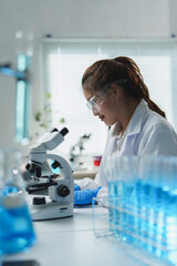 Young female scientist wearing lab coat and protective glasses using microscope while analyzing...