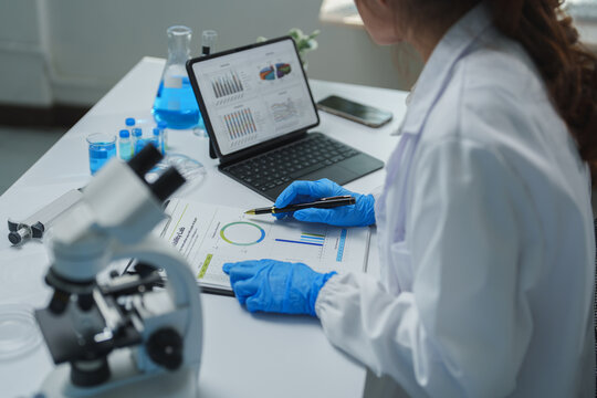 Scientist wearing blue gloves pointing at charts and graphs on a paper document while analyzing medical research data using a tablet in a laboratory setting, with a microscope and beakers nearby