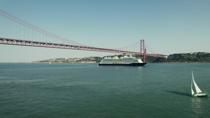 The ship sails under a red suspension bridge on a wide river in Portugal. Sailing boats on the river. Drone video.