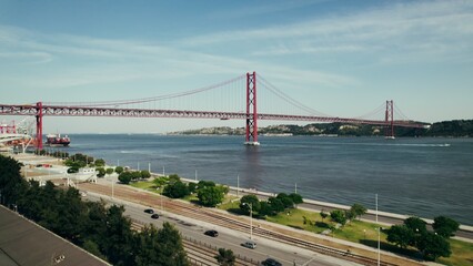 Panoramic bird's-eye view of a wide river with a red suspension bridge connecting the banks. The drone is flying over the embankment