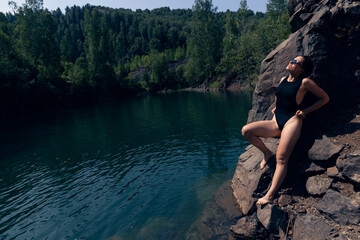 Fototapeta premium Woman in Black Swimsuit Posing on Rocky Lakeside with Forested Hills in Summer Sun