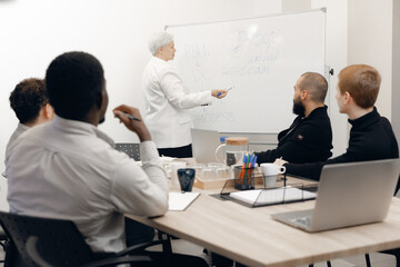 Diverse colleagues engage in business strategy meeting, analyzing plans on whiteboard. professional setting with focus on teamwork, communication, and organizational success