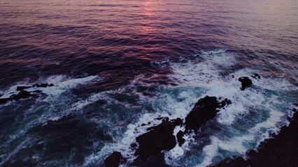 A bird's-eye view of the sea at sunset. Foam waves wash the stone reefs on the shore. Drone video