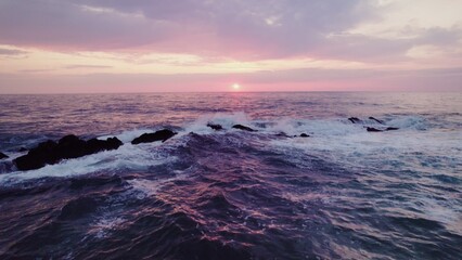 Obraz premium A bird's-eye view of the sea at sunset. Foam waves wash the stone reefs on the shore. Drone video