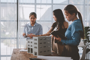 Three female engineers are evaluating a building model with integrated wind turbines, discussing...
