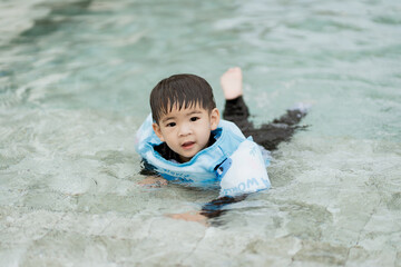 young boy wearing blue life jacket enjoys swimming in clear pool, smiling with joy. water is calm,...