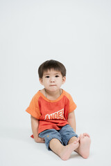 young child in red shirt and blue pants sits on floor against plain background, looking directly at camera with calm expression