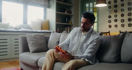 A young bearded man is intently playing on a mobile phone while sitting on a sofa in a stylish home...