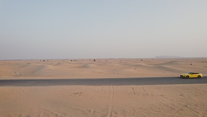 The drone flies parallel to a yellow car driving along the road among the desert sands