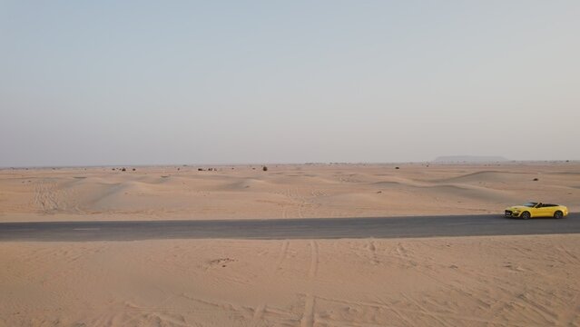 The drone flies parallel to a yellow car driving along the road among the desert sands