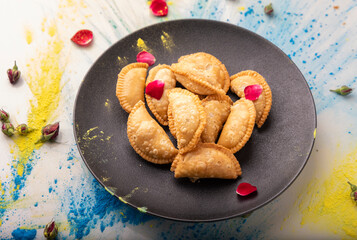 A festive presentation of Gujiya, a popular Indian sweet, placed on a black plate with vibrant Holi colors and rose petals