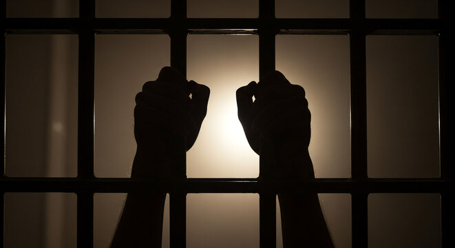 Silhouetted hands gripping prison bars with light shining through. The image is perfect for news about arrests, detention, or crimes