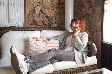 Obraz premium Happy woman relaxing on sofa, talking on mobile phone in rustic living room