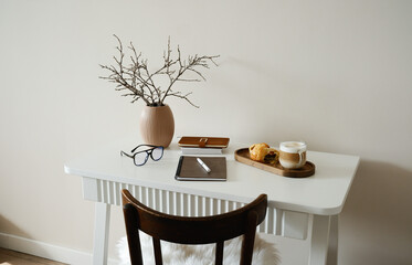 A cozy, minimalistic workspace with charming ceramic vase, dry branches, paper Easter eggs, ceramic rabbit,notebooks, glasses coffee and croissants on wooden stand.