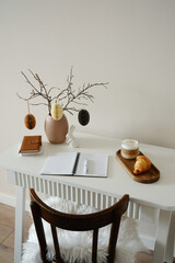 A cozy, minimalistic workspace with charming ceramic vase, dry branches, paper Easter eggs, ceramic rabbit,notebooks, opening notebooks with pen, coffee and croissants on wooden stand.