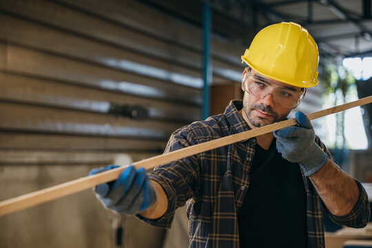Construction worker in a yellow hardhat and safety glasses inspecting a piece of wood. Ideal for carpentry, woodworking, and quality control, highlighting craftsmanship in the workshop. labor day