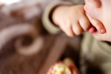 Young child in close-up licking a finger. Holding a peeled banana, showing a small portion. The background is softly blurred, highlighting the focus on the child's hand and mouth area.