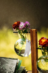 A glass vase with summer flowers, including marigolds and purple blooms, sits on a wooden structure. Water droplets are visible on the vase. A blank tag hangs from the vase, providing space for text. 