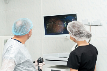 Doctor performing colonoscopy on patient on operating table. One is handling an endoscopic device while observing a monitor displaying an internal organ image, likely during a medical procedure.
