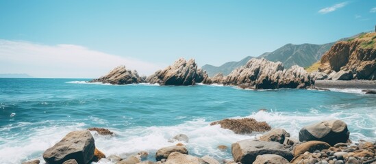 Scenic coastal landscape featuring turquoise ocean waves crashing against rocky outcrops under a clear blue sky with distant mountains in view