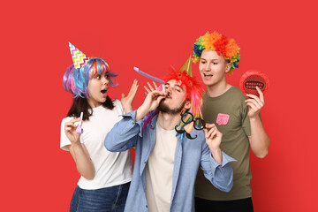 Young shocked friends in funny disguise with part whistles and whoopee cushion on red background. April fool's day celebration
