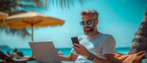 Man is sitting on a beach with a laptop and a cell phone in his hands. He is smiling and he is enjoying his time
