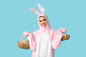 Beautiful young woman in bunny costume holding wicker baskets with Easter cake and eggs on blue background