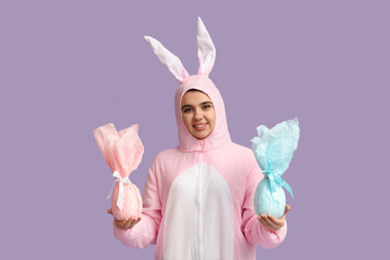 Happy young woman in bunny costume with Easter gift eggs on purple background
