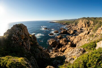 Obraz premium Rocky coastline with crashing waves and vibrant green vegetation in sunny weather