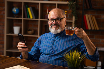 Retired Indian senior man reads news and browses internet on smartphone while sipping black coffee