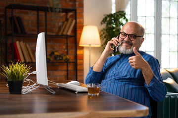 Indian elderly man in kurta works on desktop, talks on the phone, and drinks coffee at home