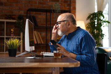 Indian elderly man in kurta works on desktop, talks on the phone, and drinks coffee at home
